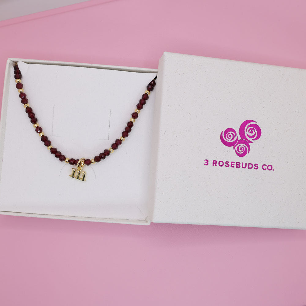 Necklace with a gold charm on a box labeled '3 Rosebuds Co.' against a pink background