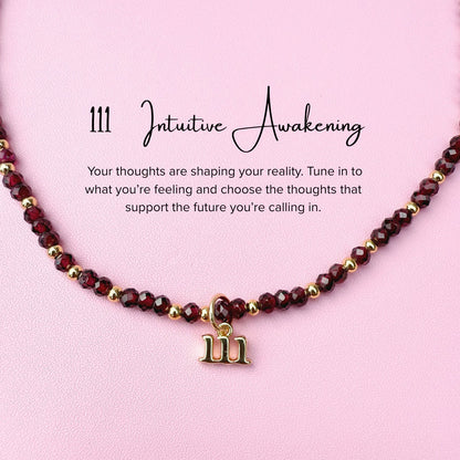Necklace featuring maroon garnet beads and a gold 111 angel number charm on a pink background, accompanied by text about intuitive awakening.