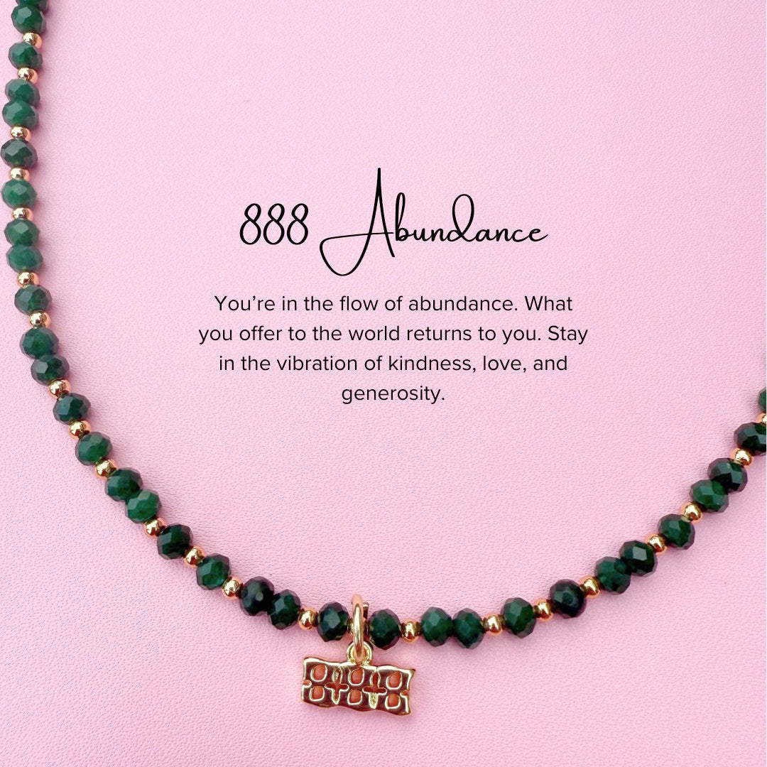 Necklace with green beads and a 888 angel numer gold charm on a pink background with text about abundance and positivity.
