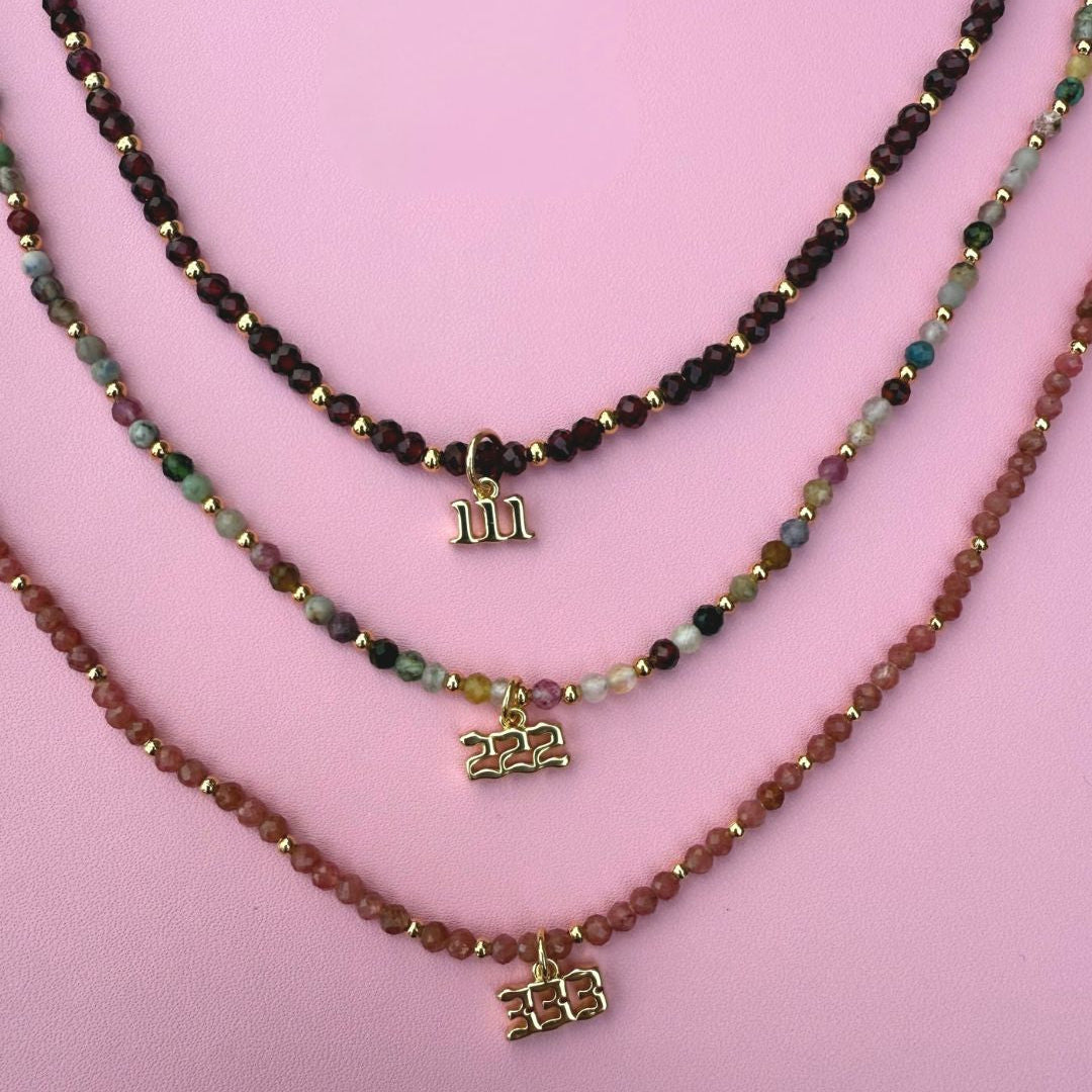 Three beaded crystal necklaces with angel number charms 111, 222, 333 on a pink background