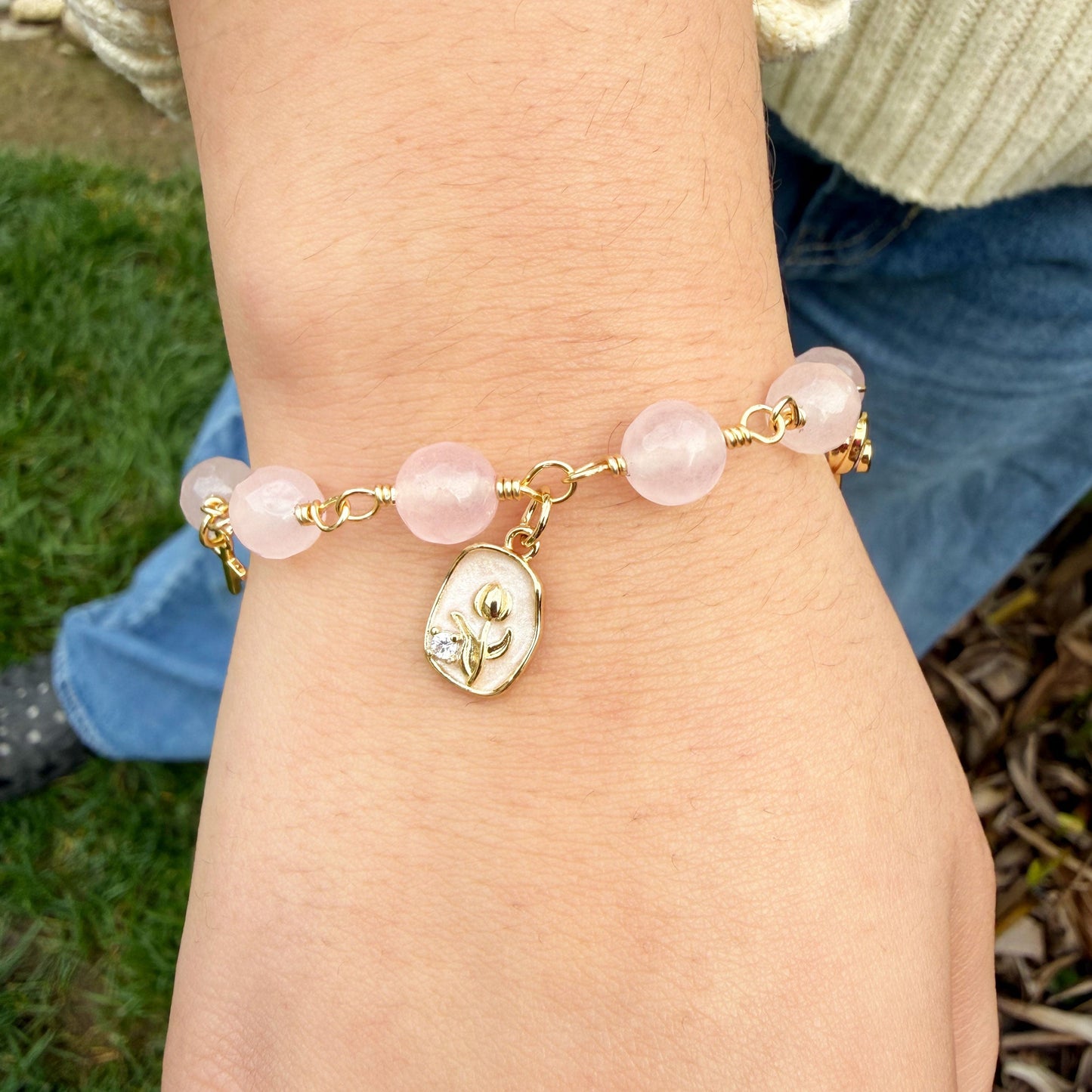 Close-up of a wrist wearing a rose quartz rosary bracelet with pink beads and a gold charm, outdoors.